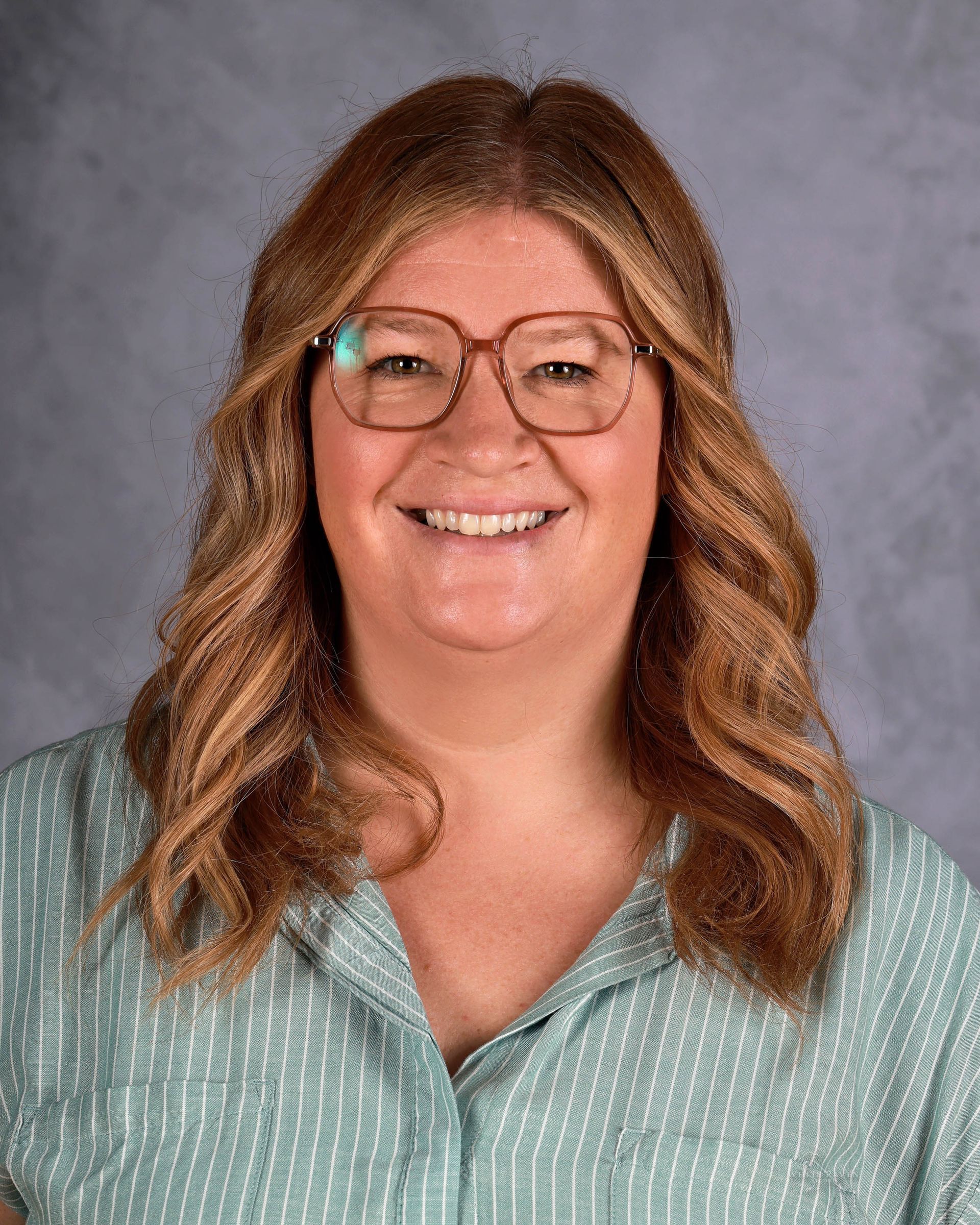 Smiling woman with long wavy brown hair, wearing glasses and a striped button-down shirt.