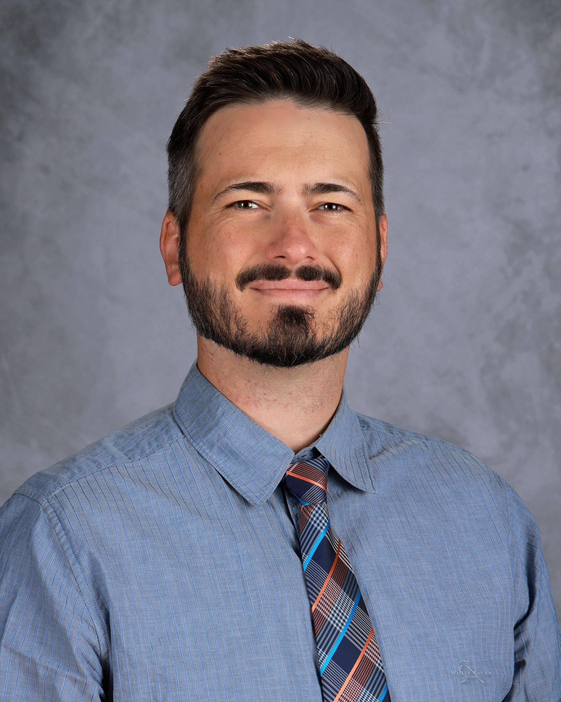 Man with brown hair and beard, wearing a blue shirt and patterned tie, smiling against a gray background.