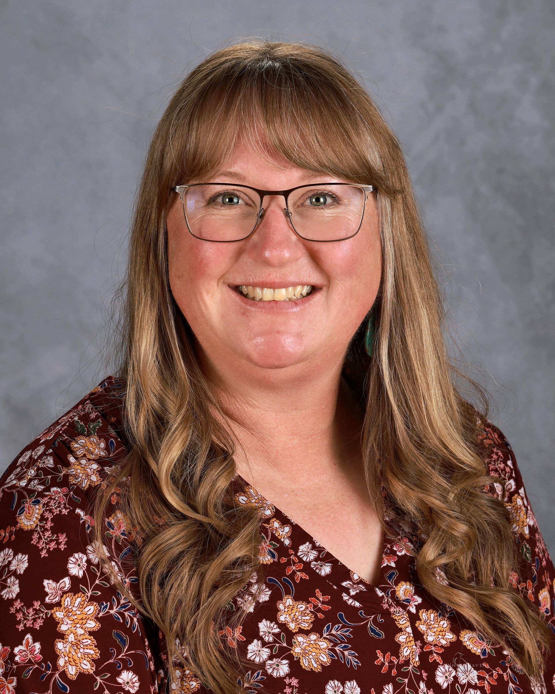 Woman with glasses, long hair, and floral shirt smiles at the camera.