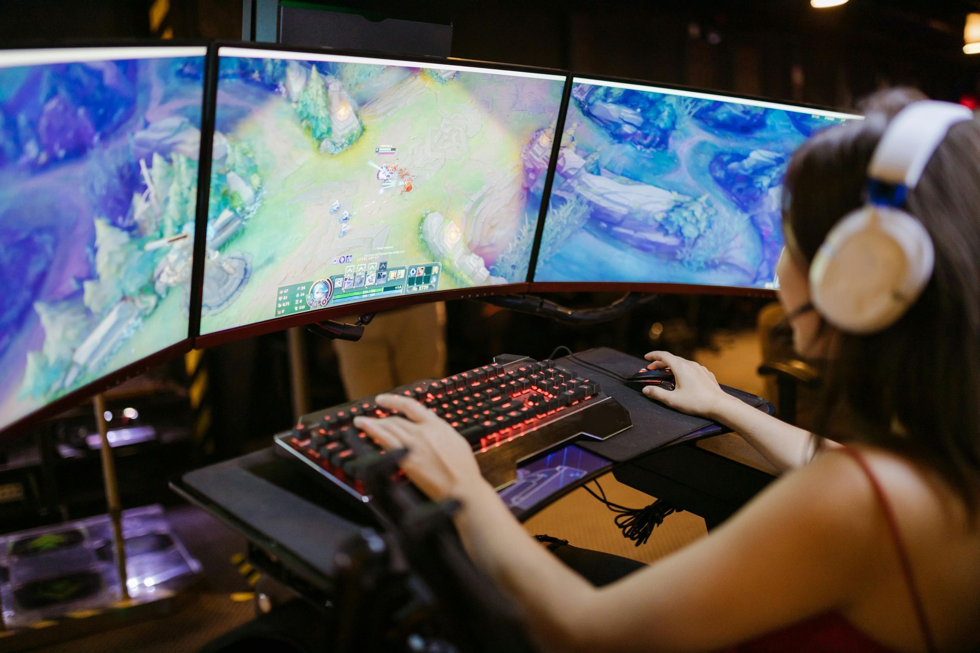 Woman gaming on three curved monitors, using keyboard and mouse, wearing headphones, indoors.