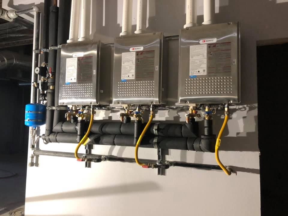 Three water heaters are lined up on a wall next to pipes.