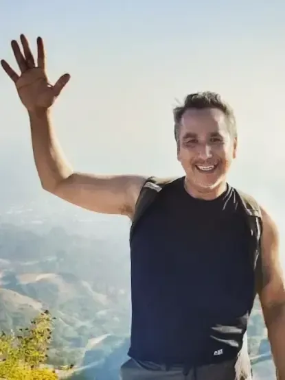 Joseph R Cashman waving with a smile against a mountain backdrop. He wears a black tank top.