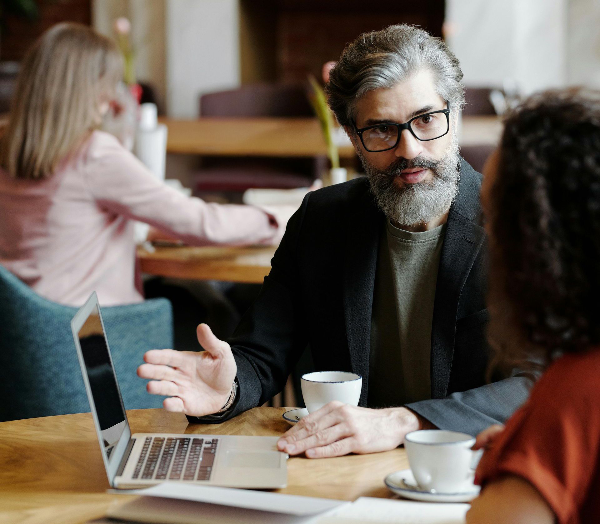 Man with beard gestures toward laptop, conversing with woman at a cafe table. Others work in the background.