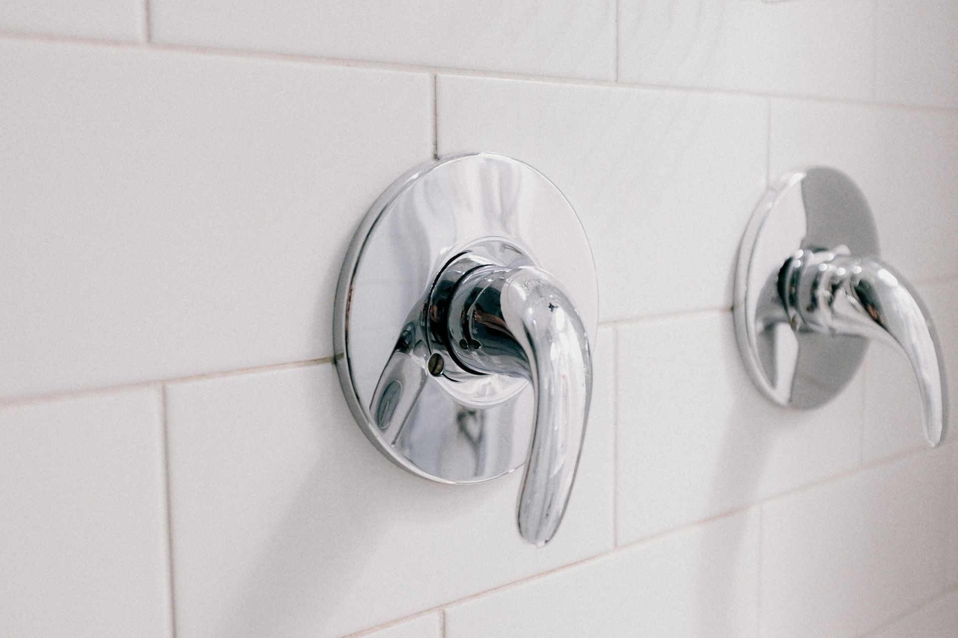 Two chrome shower handles on white tiled wall.