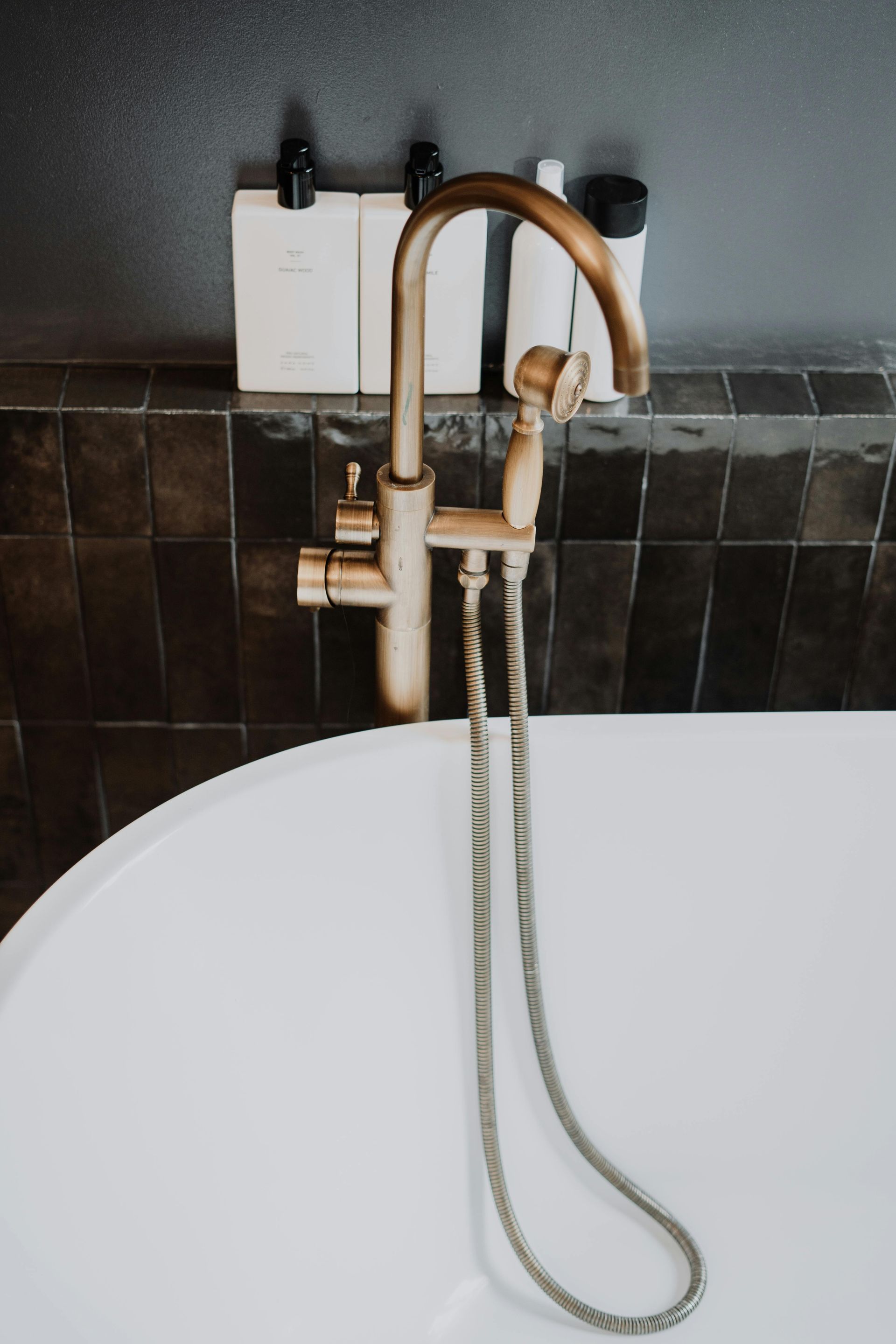 Antique bronze faucet over a white bathtub, set against dark tiled wall with toiletries.