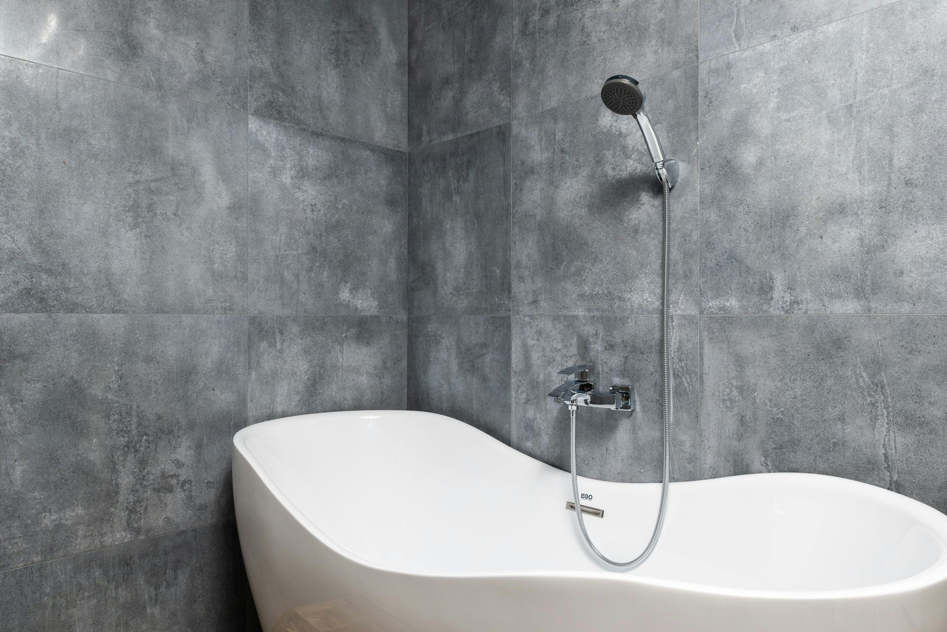 A modern white bathtub with silver shower fixtures set against gray tiled walls.