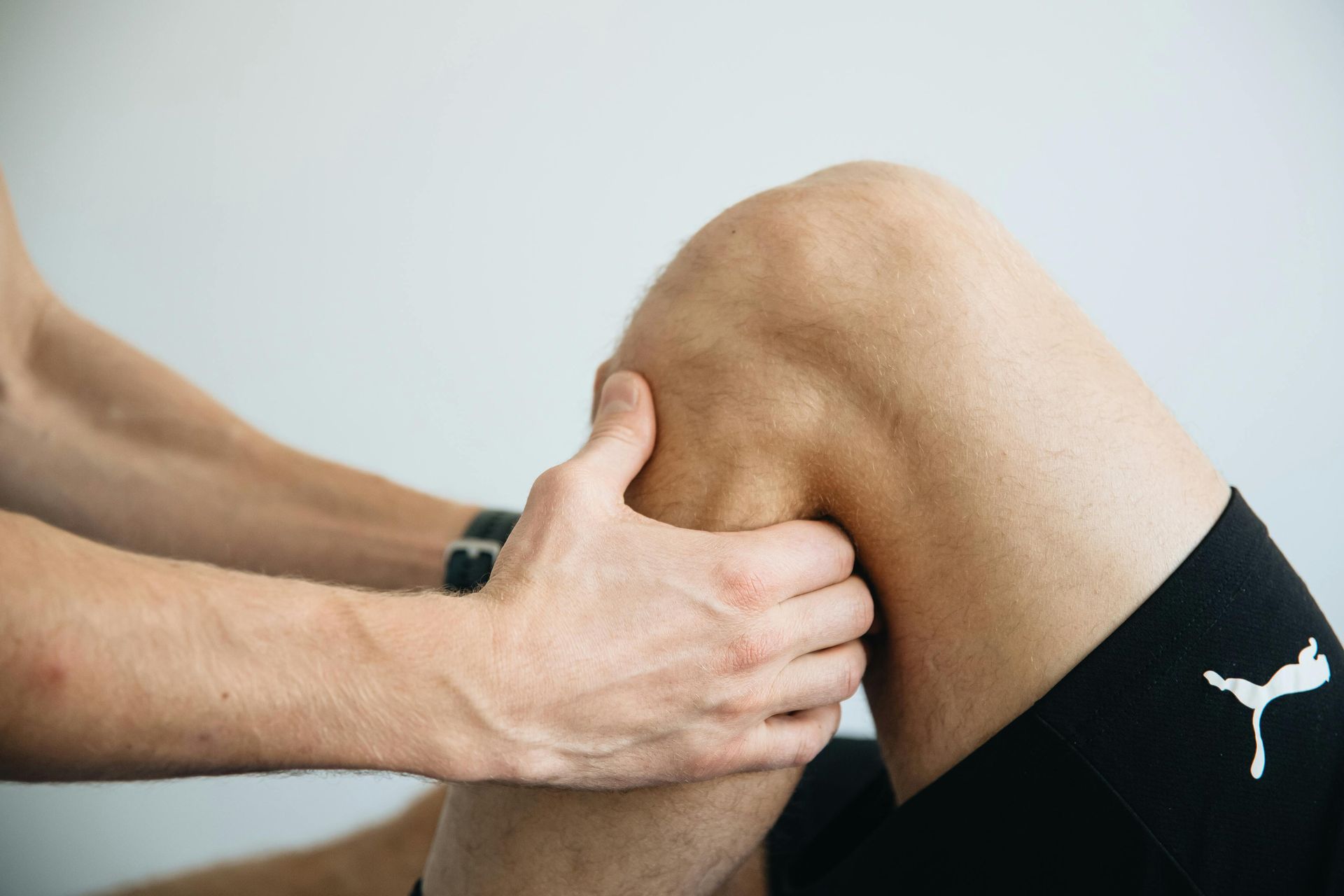 Hands examining a person's knee. Leg flexed, skin tone visible, set against a plain background.