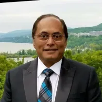 Man in suit and tie, smiling, outdoor setting with water and greenery in background.