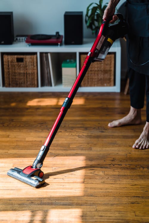 Person vacuuming hardwood floor with a red stick vacuum. Sunlight streams in.