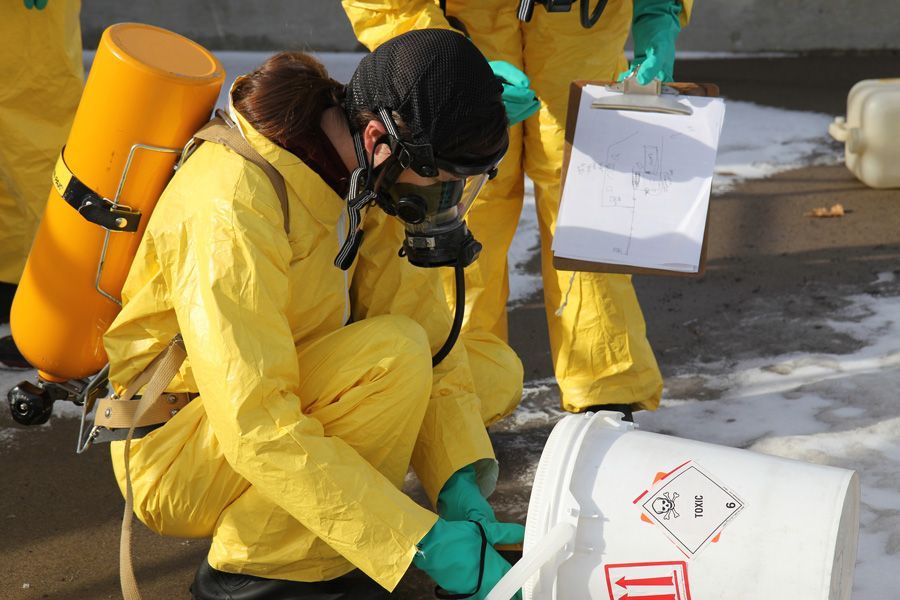 Person in yellow hazmat suit inspecting a spilled container, outdoors.