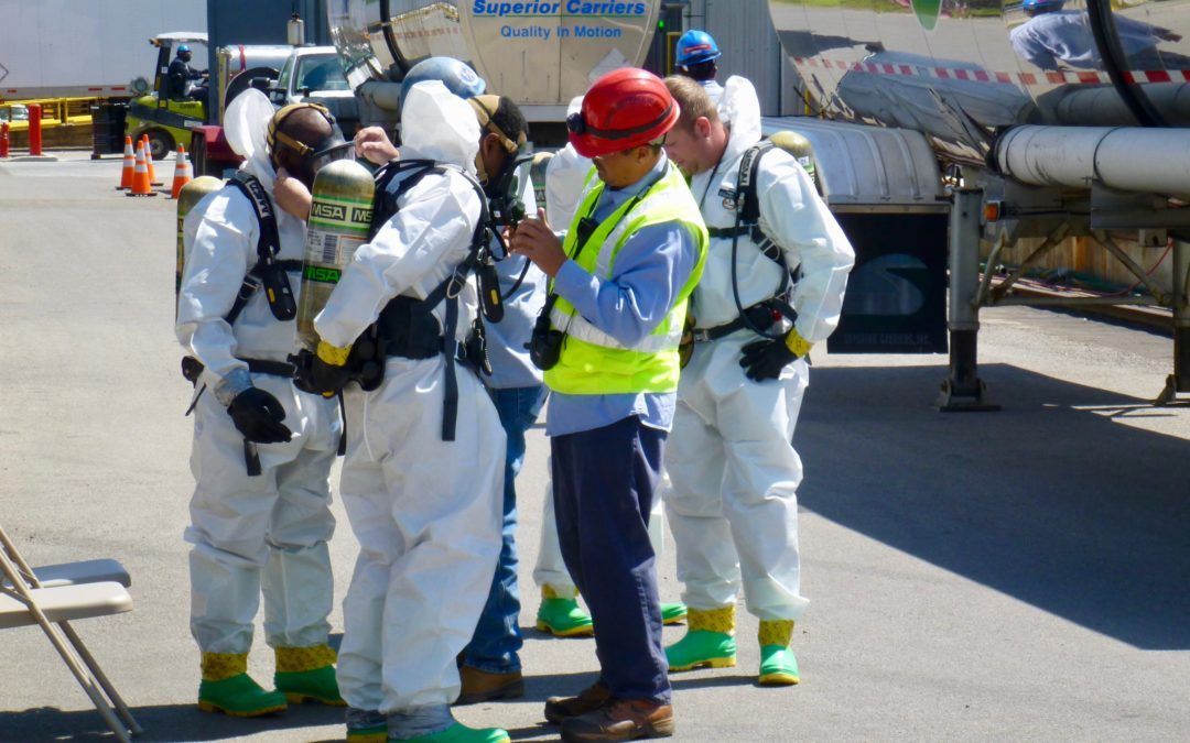 Workers in hazmat suits being briefed outdoors. One in a red hard hat, others are wearing protective gear.