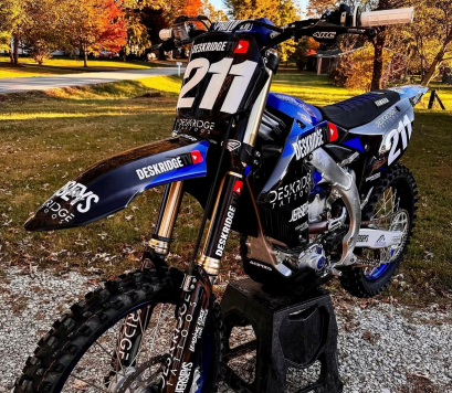Blue dirt bike with number 211 parked on gravel in an autumn outdoor setting with fallen leaves.
