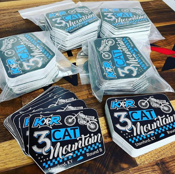 Stickers piled on a wooden table, featuring blue and black “3 Cat Mountain” designs.