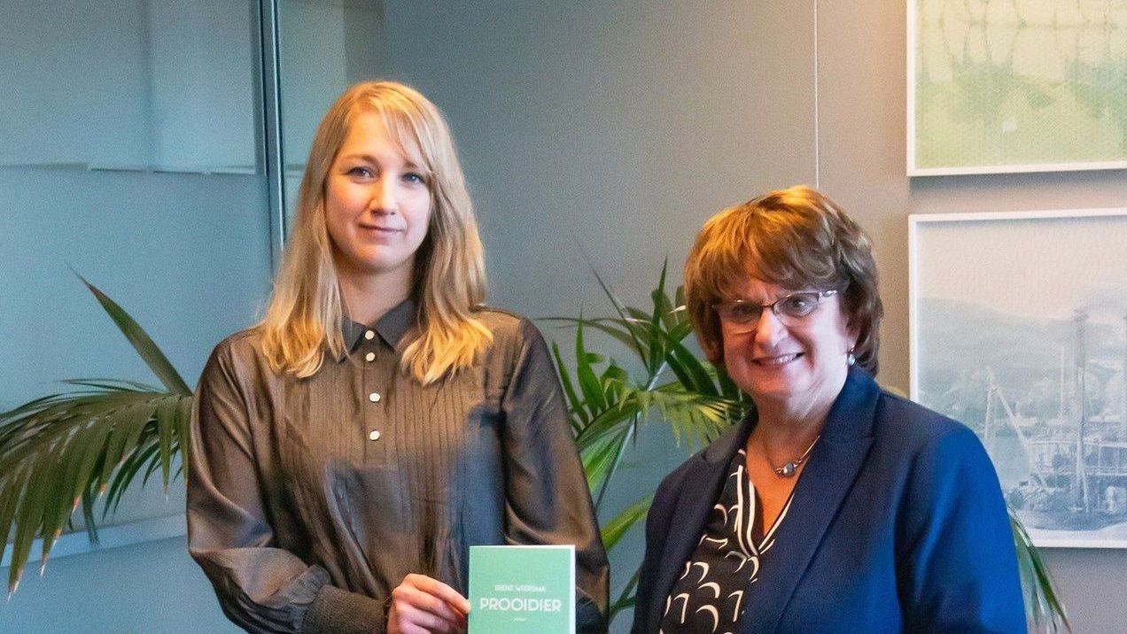 Twee mensen staan ​​binnen, houden een klein groen bordje vast en glimlachen naar de camera.