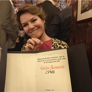 Een lachende vrouw houdt een certificaat vast tijdens een formeel evenement, met mensen en ingelijste kunst op de achtergrond.