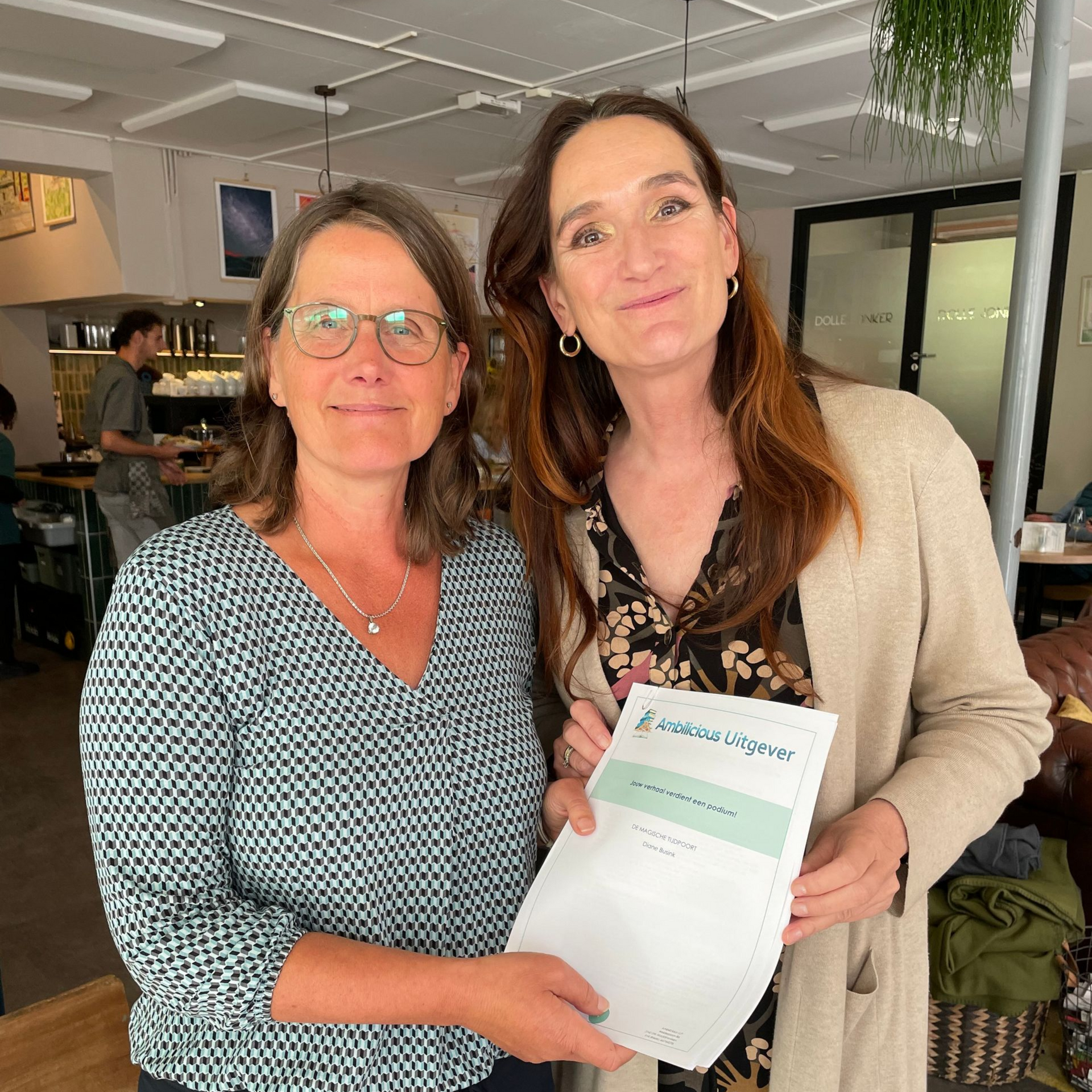 Twee vrouwen staan ​​binnen, glimlachend en met een certificaat en een papieren boekje in hun handen.