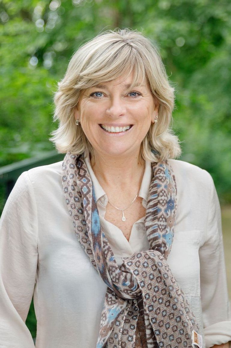 Lachende vrouw in een lichte blouse en een sjaal met patroon, buiten tegen een wazige groene achtergrond.