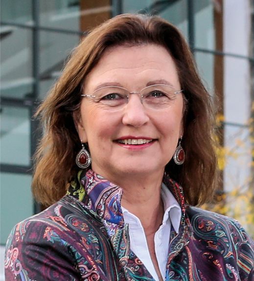 Lachende vrouw met bril en kleurrijke jas staat buiten voor een gebouw.