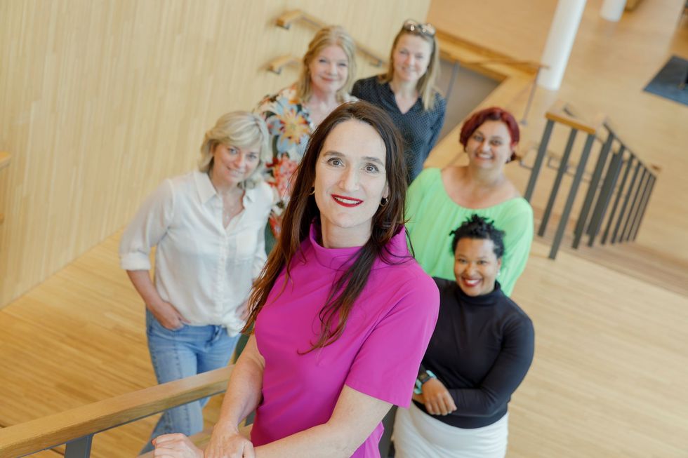 Een groep van vijf mensen staat lachend op een houten trap, aangevoerd door een vrouw in een roze top.