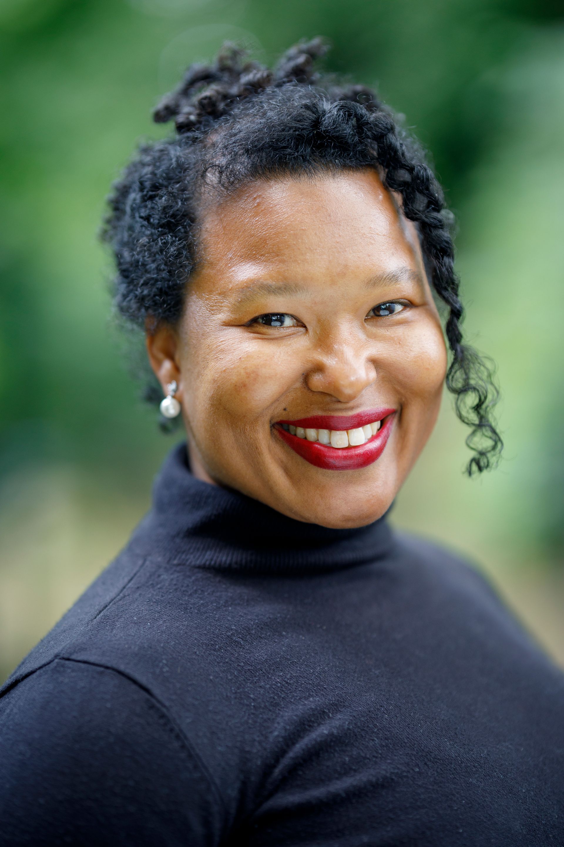 Een lachend persoon in een zwarte coltrui tegen een wazige groene achtergrond.