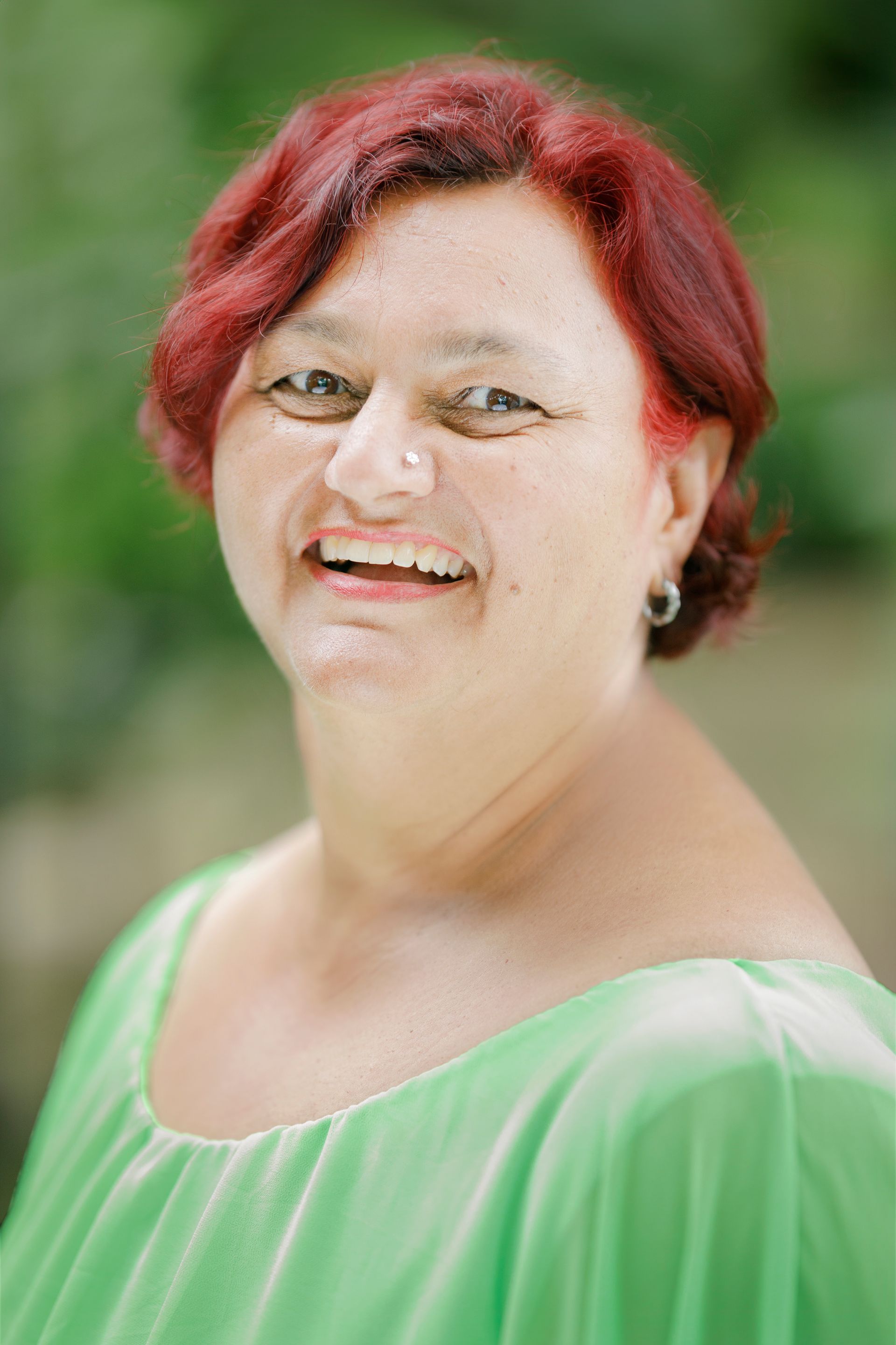 Een lachend persoon met kort rood haar en een felgroen topje, buiten met een wazige groene achtergrond.