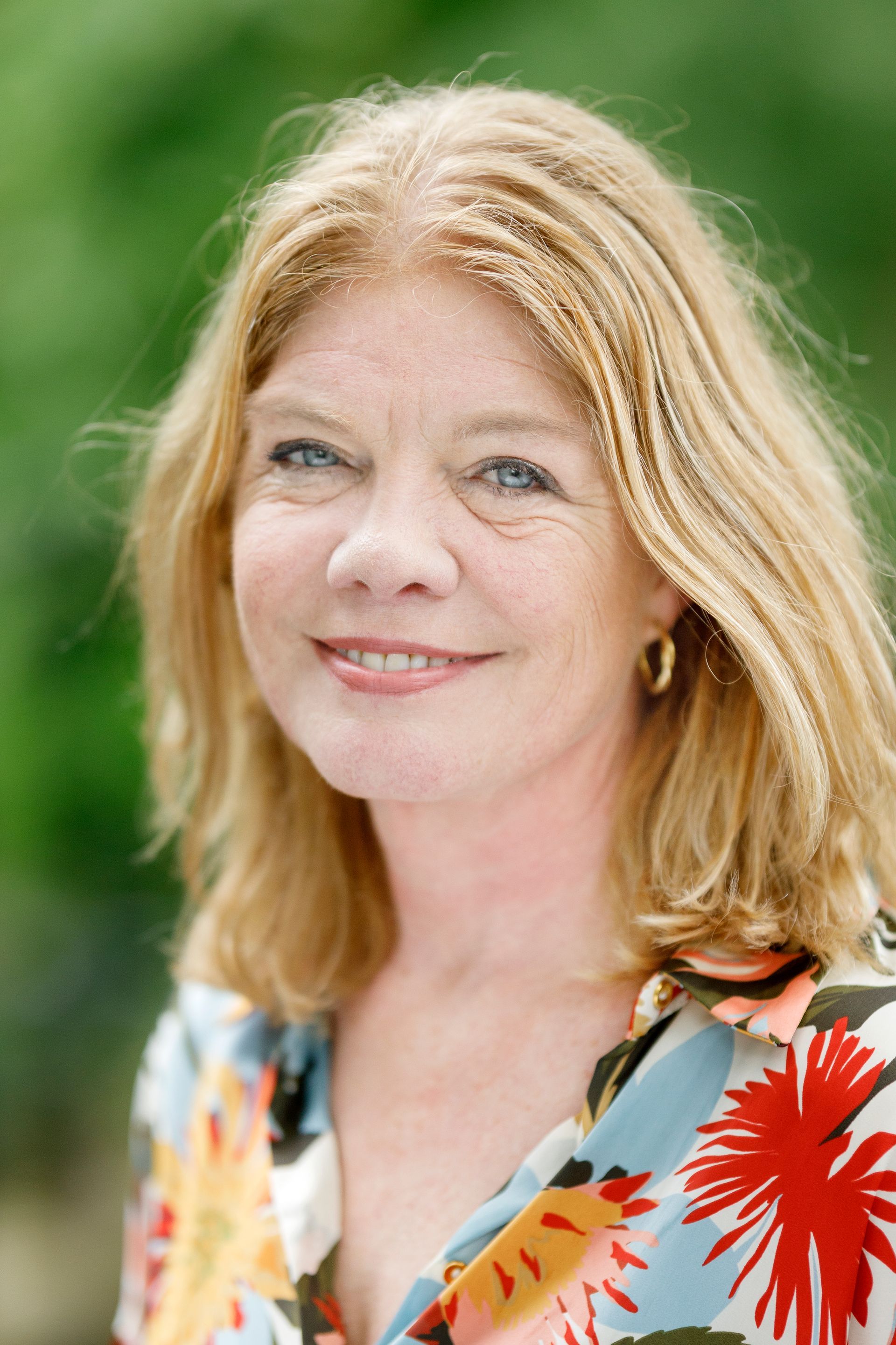 Lachende vrouw met blond haar, gekleed in een kleurrijke top met bloemenprint, tegen een groene achtergrond.