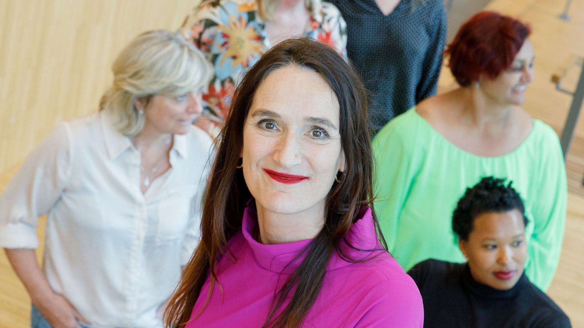Een groep mensen staat binnen bij elkaar, met een vrouw in een felroze top in het midden vooraan.