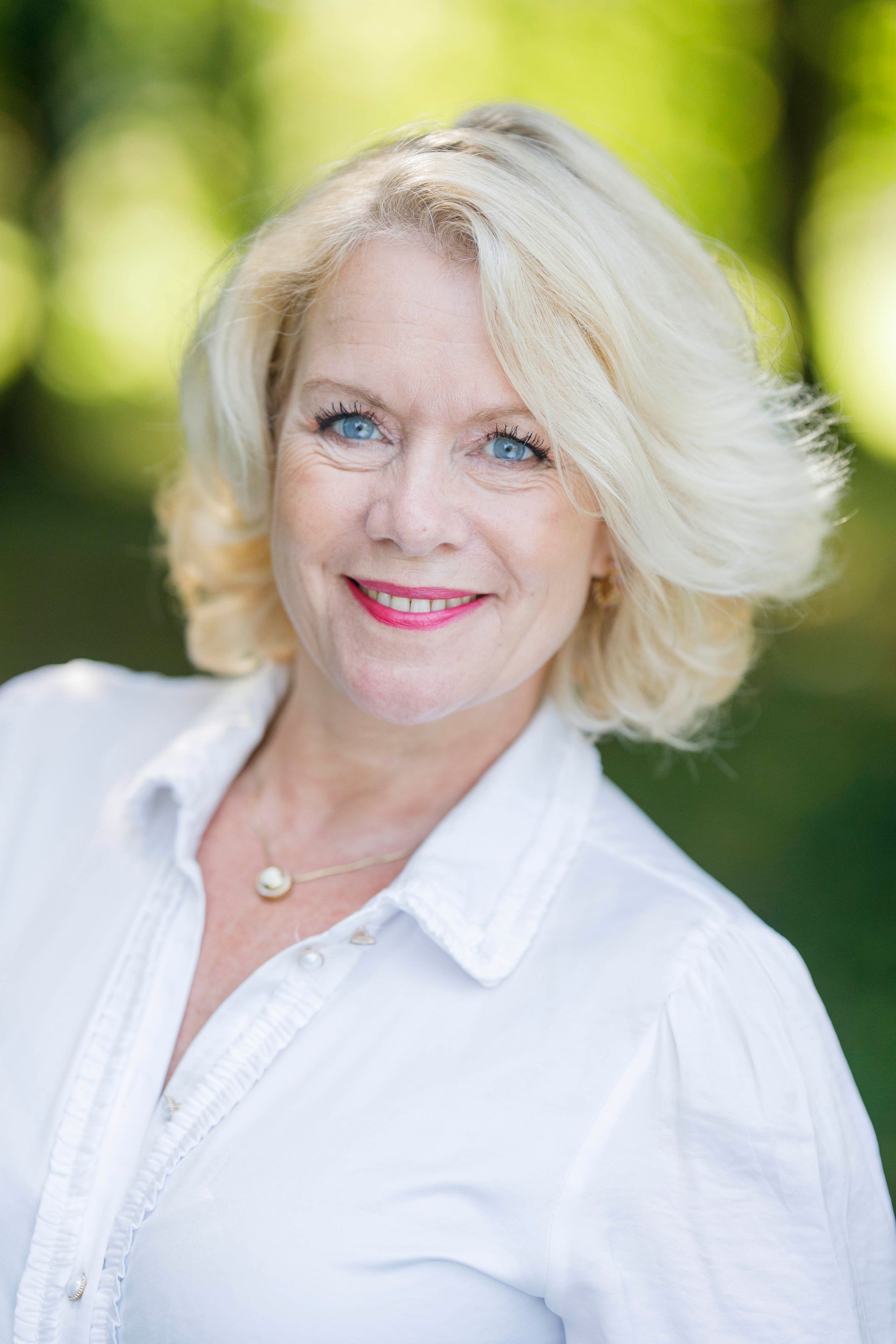 Lachende blonde vrouw in een witte blouse tegen een wazige groene buitenachtergrond.
