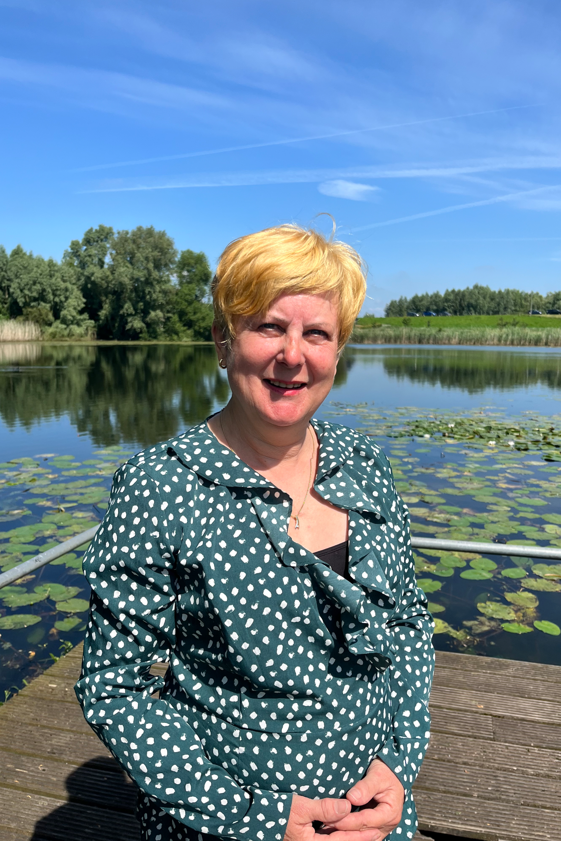 Vrouw in een groene blouse met stippen zit aan een meer met waterlelies onder een blauwe hemel.
