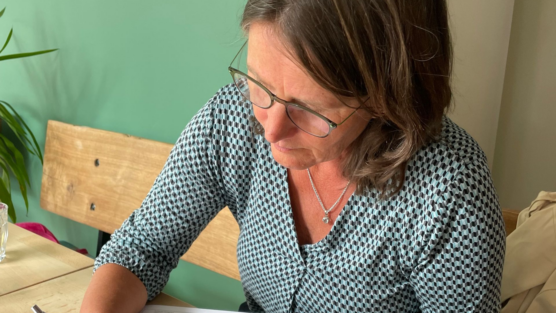 Vrouw met bril buigt zich over een tafel en schrijft in een lichte kamer met een groene muur.