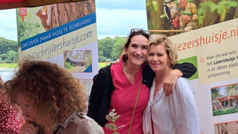 Twee lachende vrouwen poseren voor een spandoek bij een kraam op een buitenmarkt.