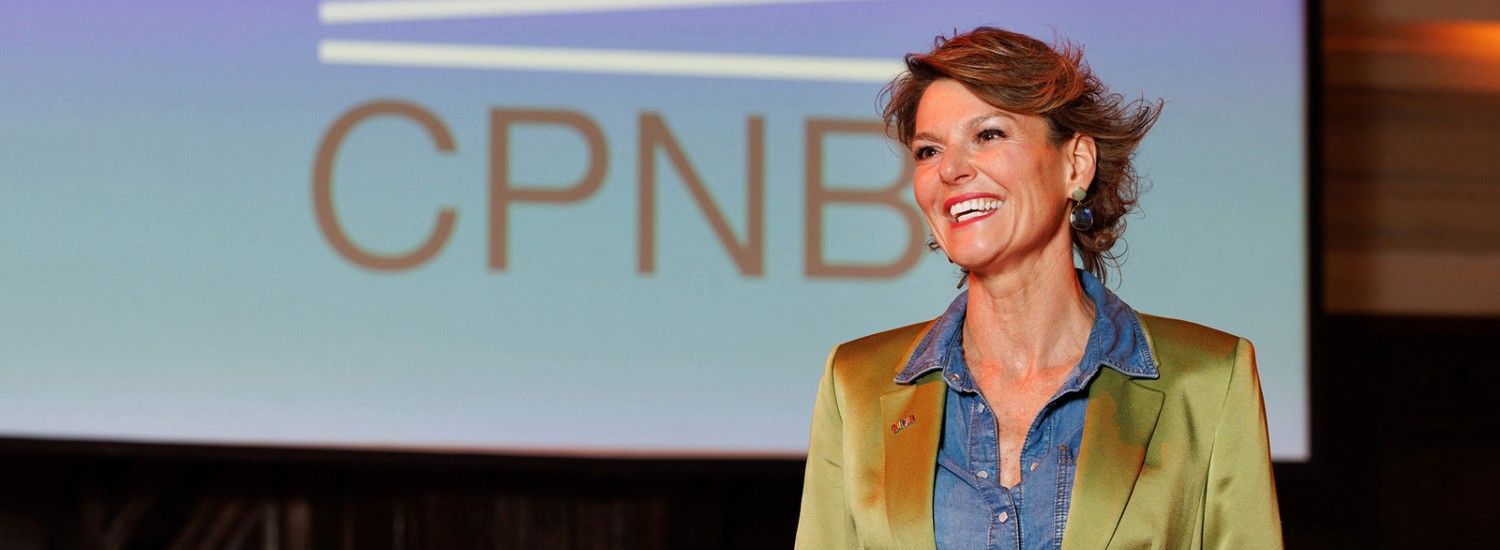 Vrouw lachend op het podium in een groene blazer voor een CPNB-achtergrond.