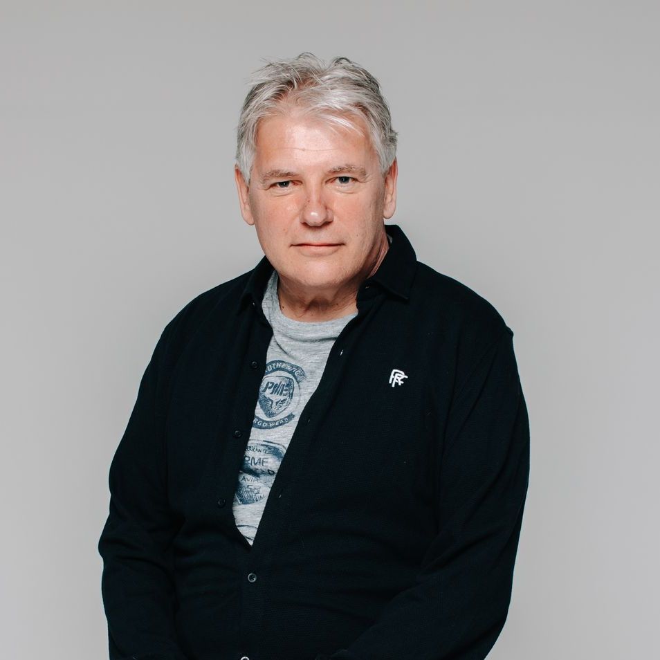 Portret van een persoon in een zwarte jas over een grijs shirt, zittend tegen een lichtgrijze achtergrond.