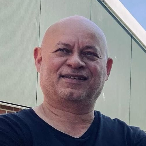 Een lachende, kale man in een donker shirt staat buiten bij een lichtgekleurd gebouw.