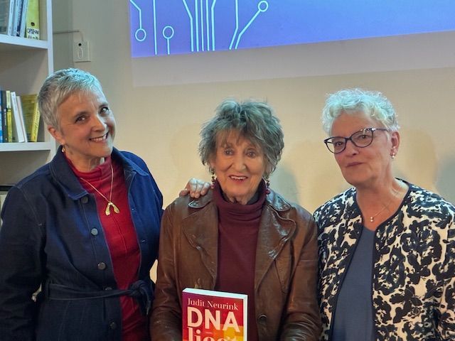 Drie vrouwen poseren binnen naast een grote DNA-boekenpresentatie en een muurgrafiek.