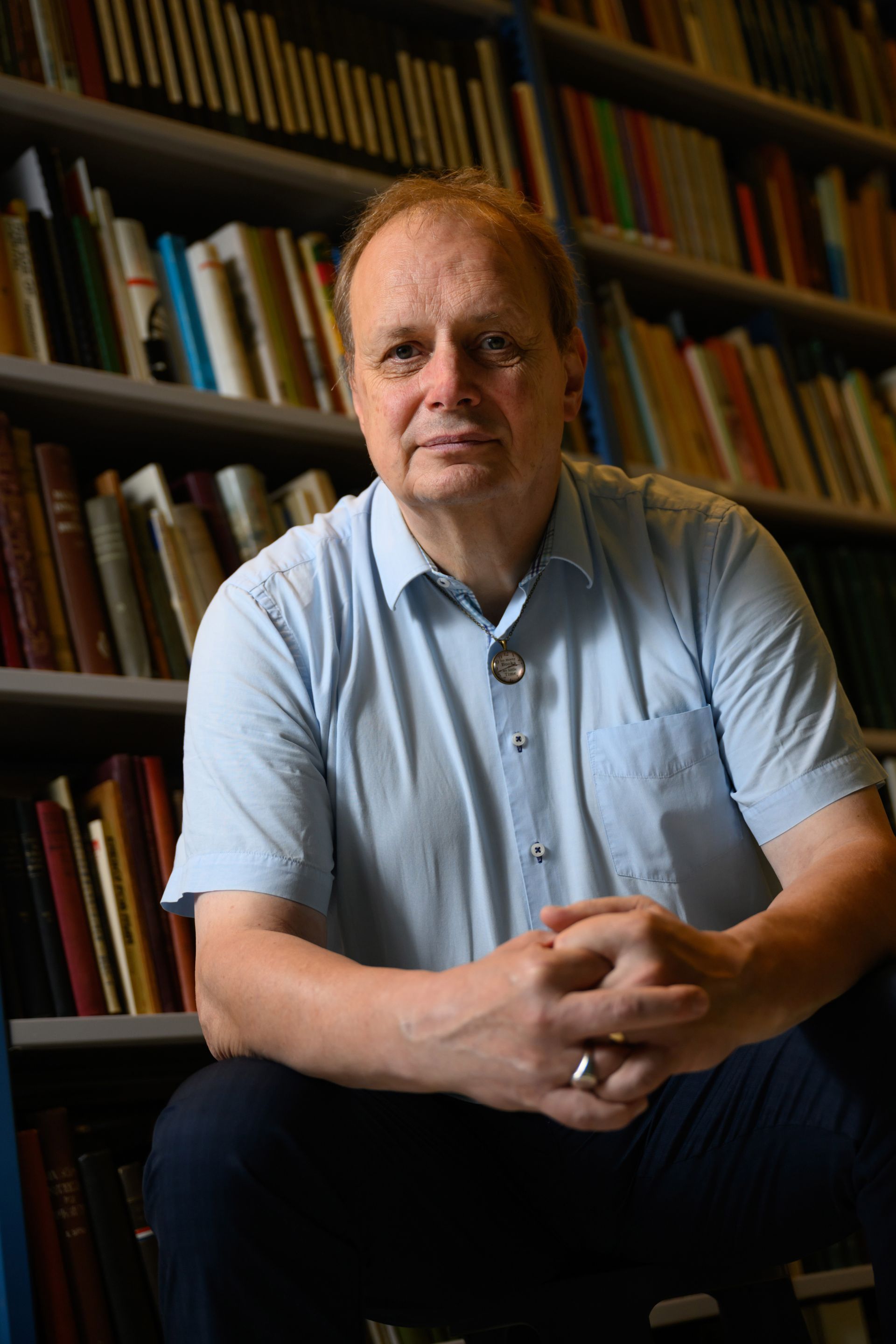 Een man zit voor een boekenkast, gekleed in een lichtblauw shirt, met zijn handen ineengevouwen, en kijkt naar de camera.