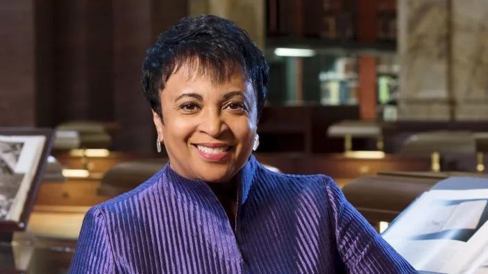 Een lachende vrouw in een paarse jas zit binnen, met boekenplanken en ingelijste foto's op de achtergrond.