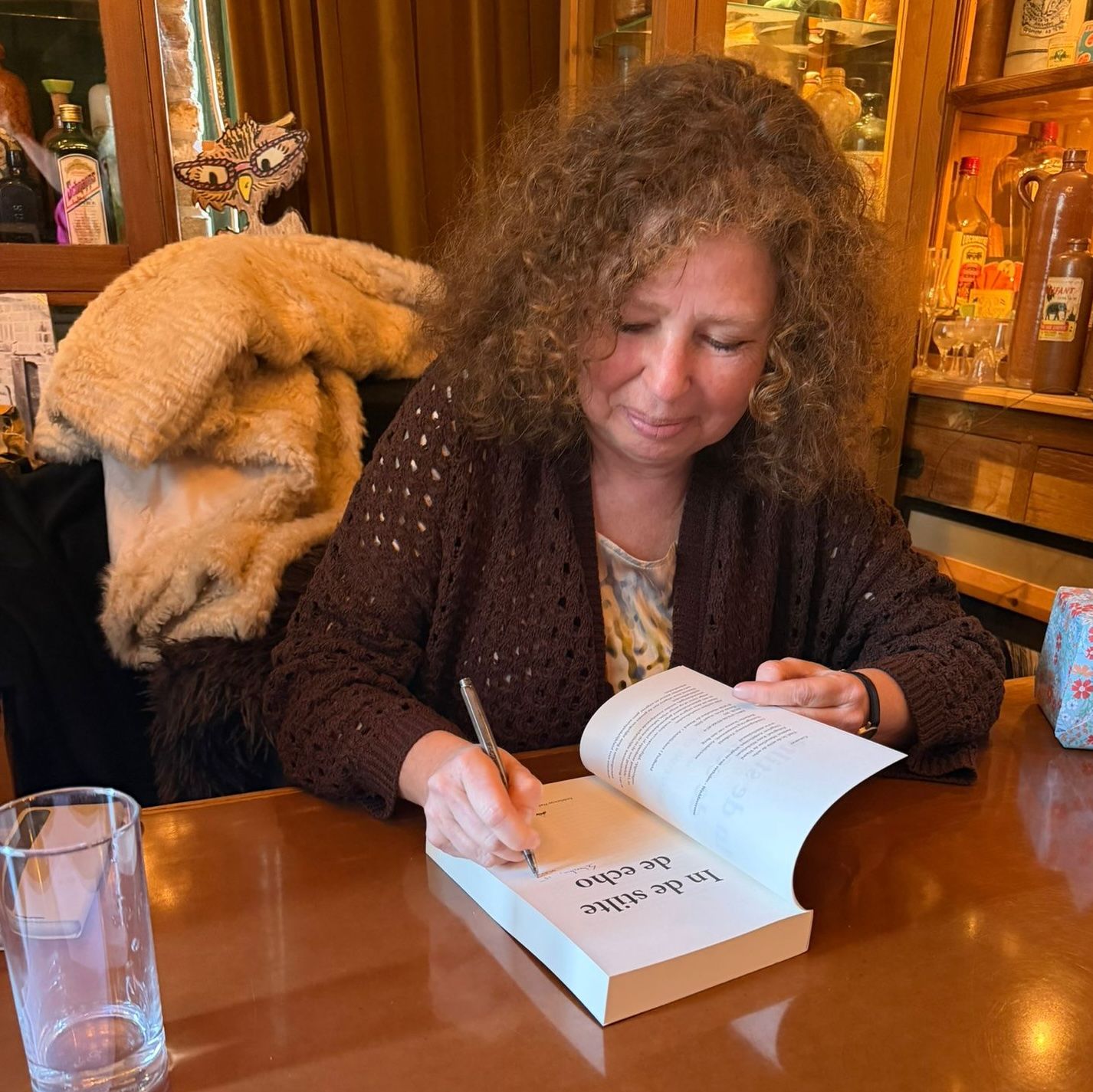 Een persoon schrijft in een boek aan een houten tafel naast een grote, lichtbruine hond in een gezellige kamer.