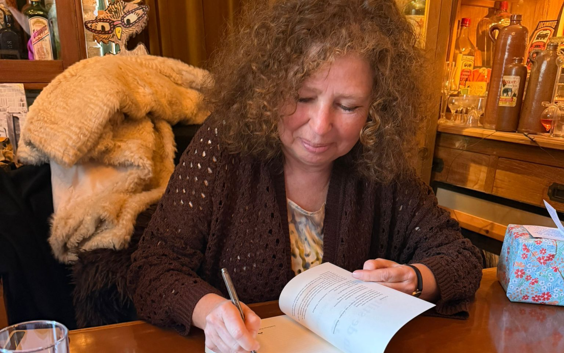 Een vrouw schrijft in een notitieboekje aan een houten tafel, met een beige jas over een stoel gedrapeerd en cadeaudozen in de buurt.