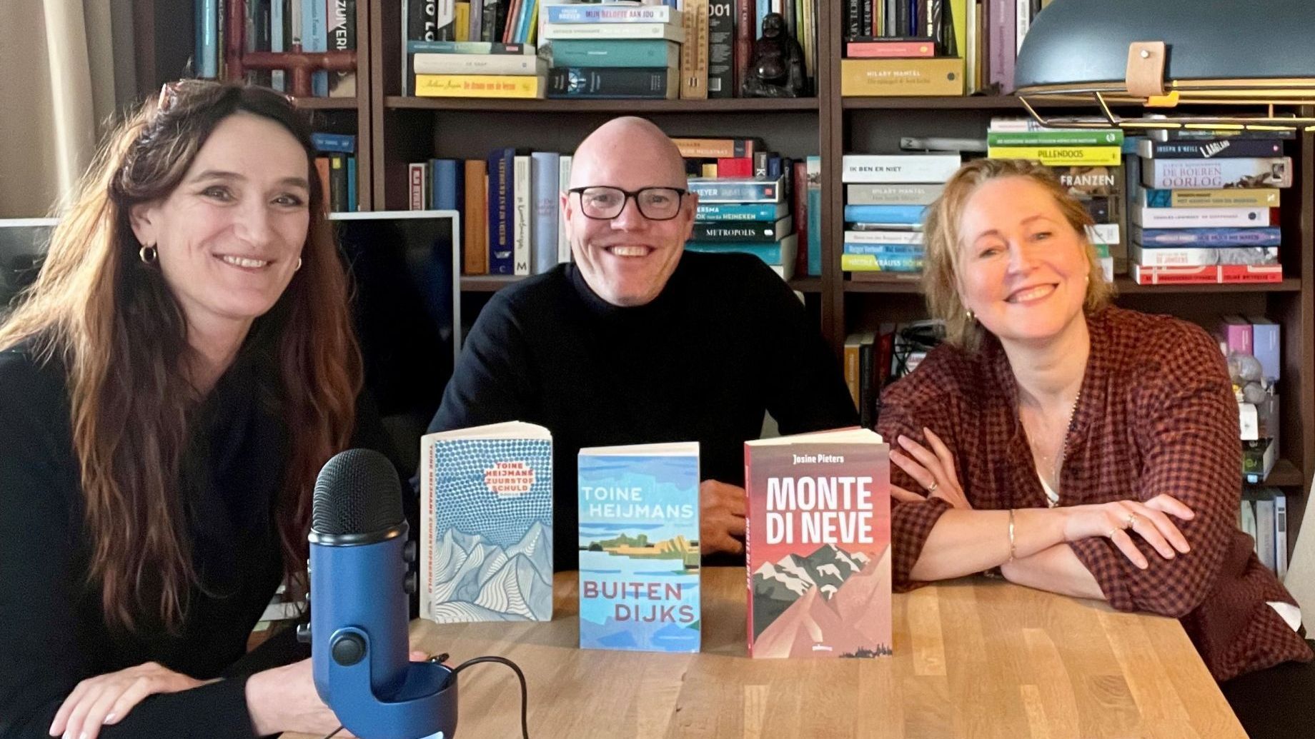 Drie mensen glimlachen aan een tafel met boeken en een blauwe microfoon voor een boekenplank.