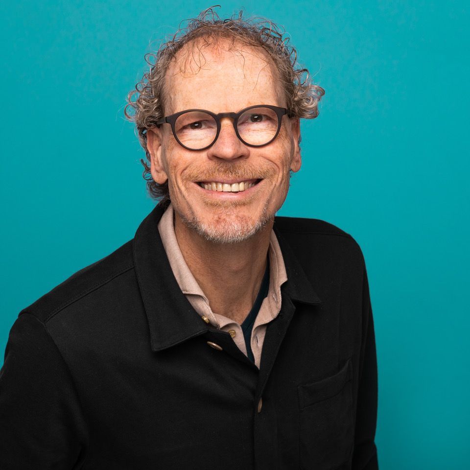 Lachende man met krullend haar en een bril in een zwarte jas tegen een turquoise achtergrond.