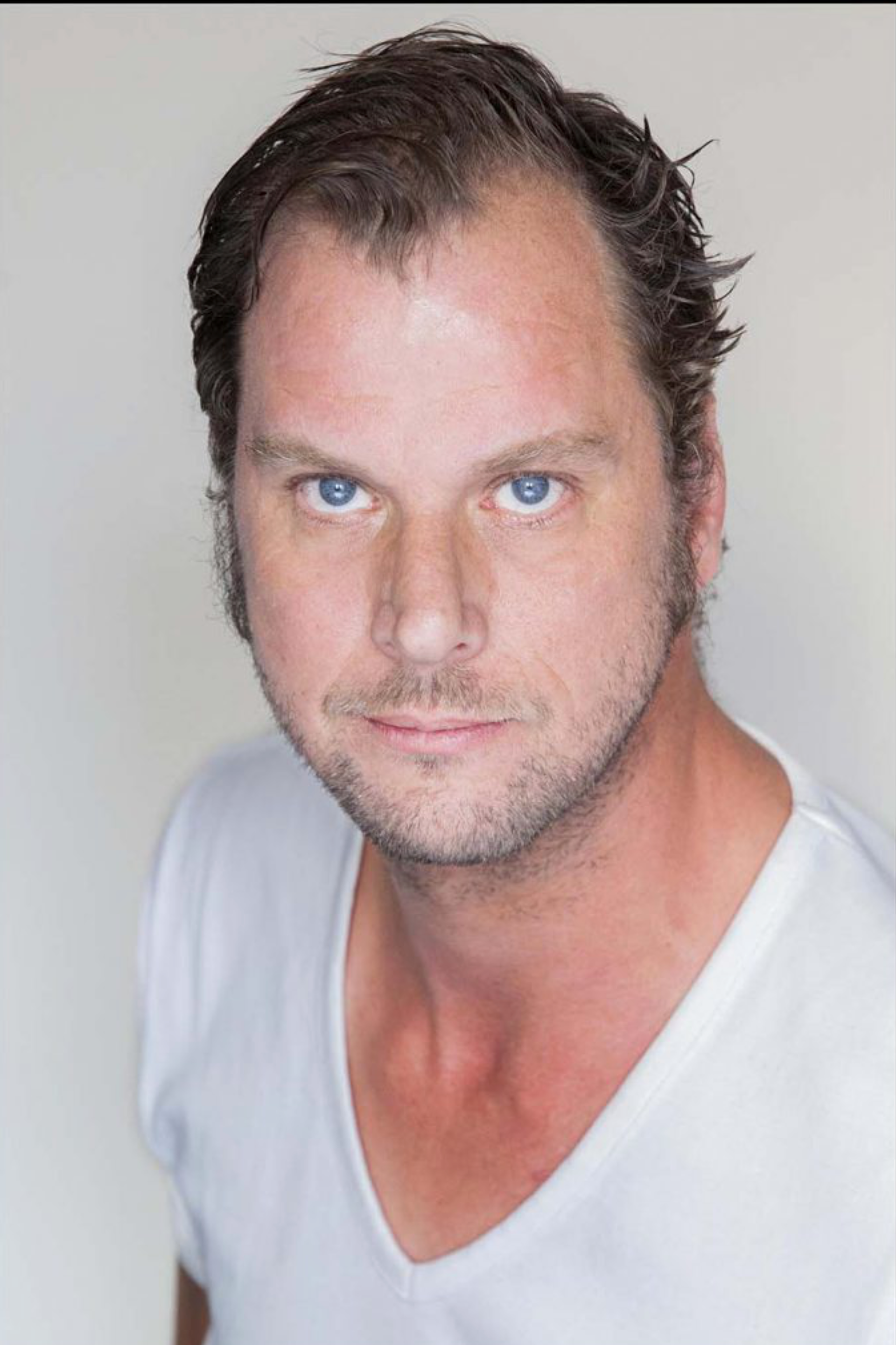 Portret van een man met kort donker haar en blauwe ogen, gekleed in een wit T-shirt met V-hals, tegen een lichte achtergrond.