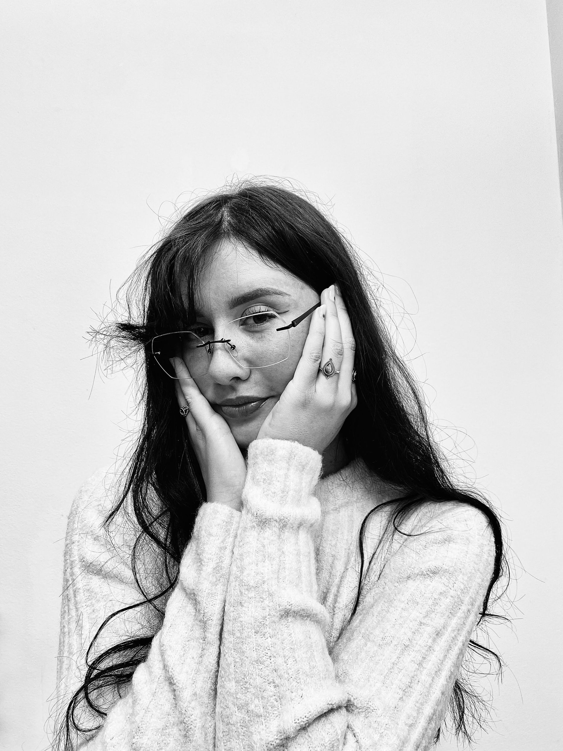 Persoon met bril en trui poseert met de handen op de wangen tegen een effen muur.