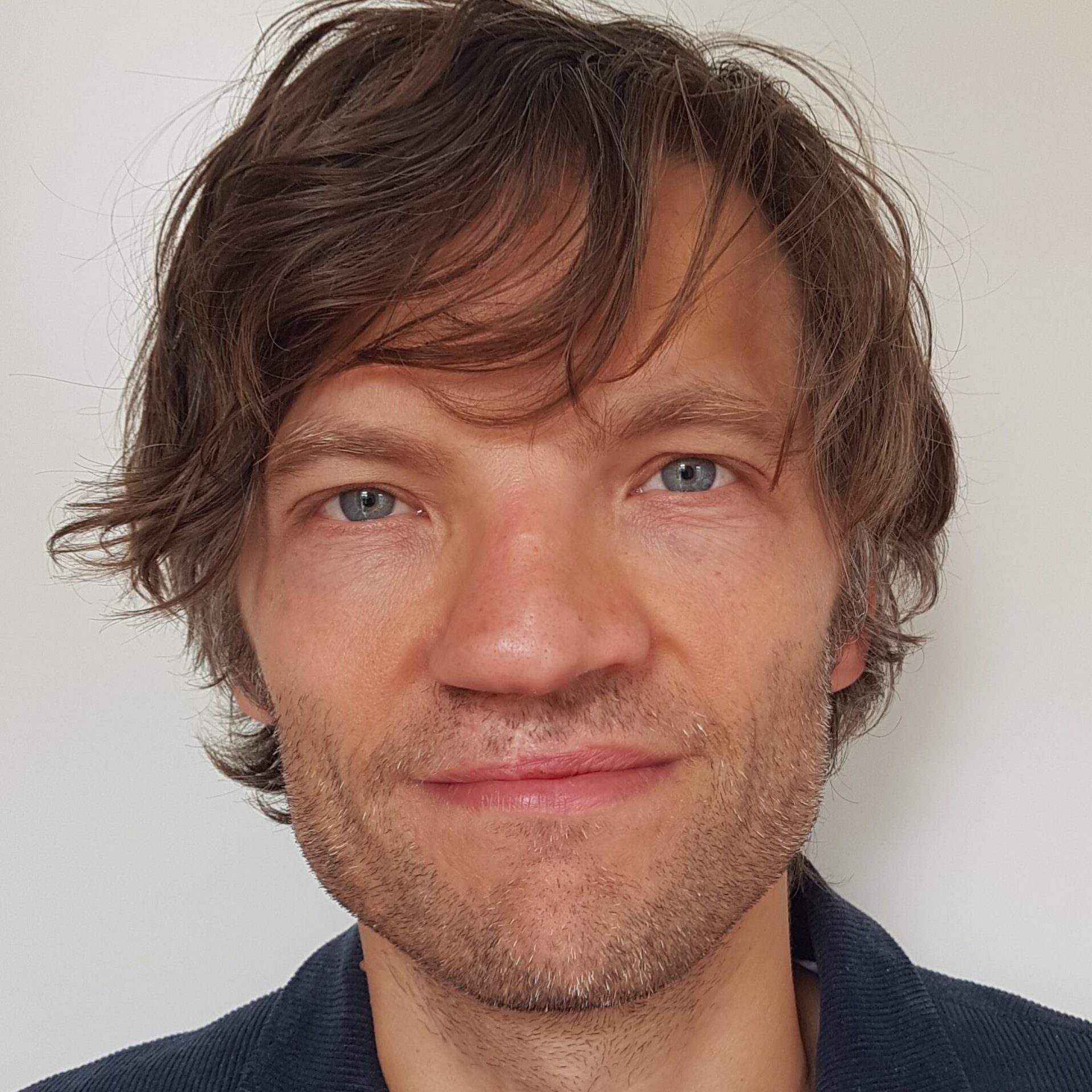 Close-up portret van een man met warrig bruin haar en blauwe ogen tegen een effen witte achtergrond.