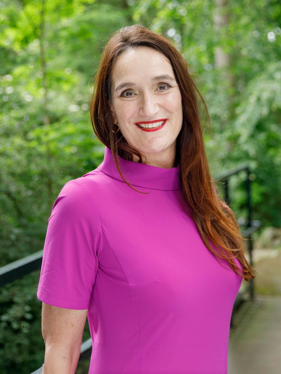 Vrouw in een felroze top staat buiten op een lommerrijk pad en lacht naar de camera.