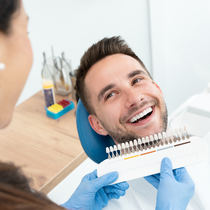 A man is smiling while sitting in a dental chair while a woman holds a tooth color chart.