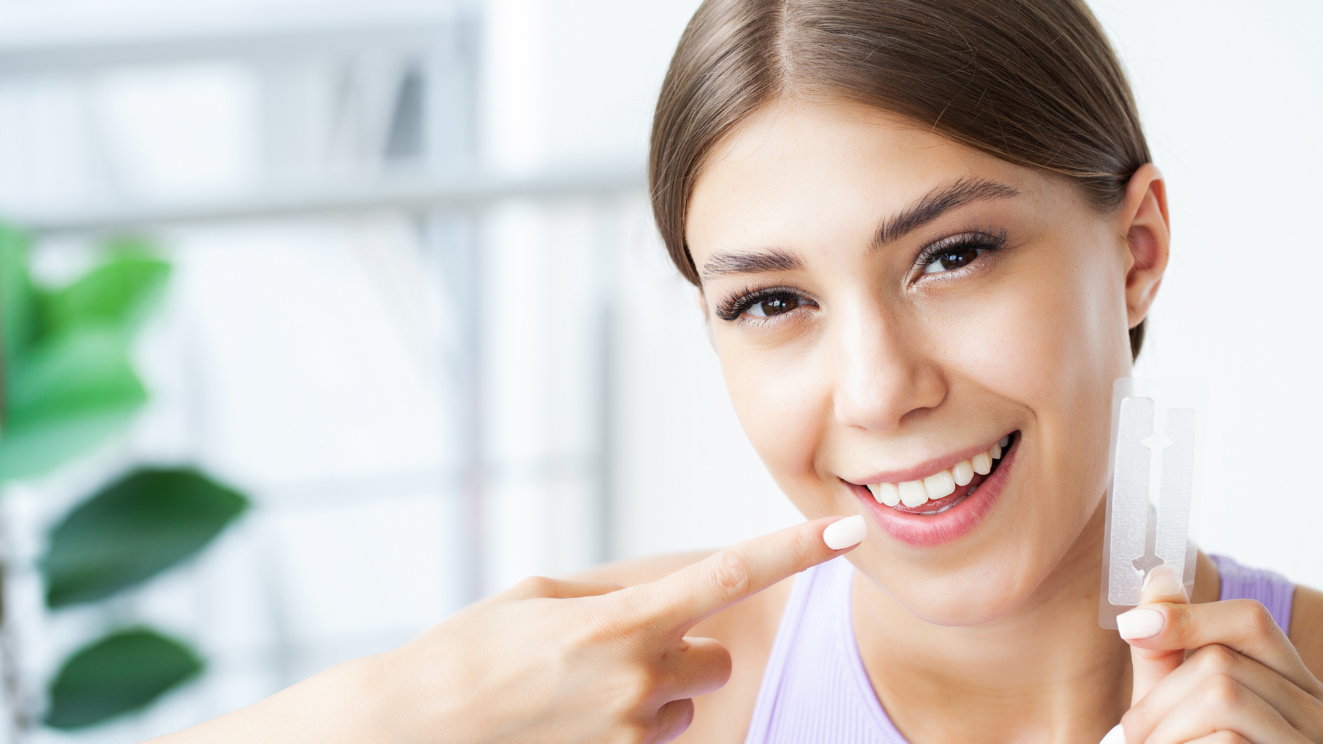 A woman is smiling and pointing at her teeth while holding a whitening strip.