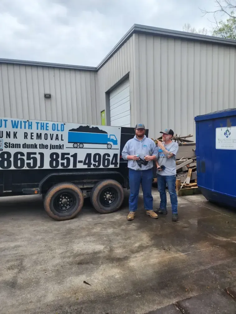 Two men stand by a junk removal trailer in front of a warehouse.
