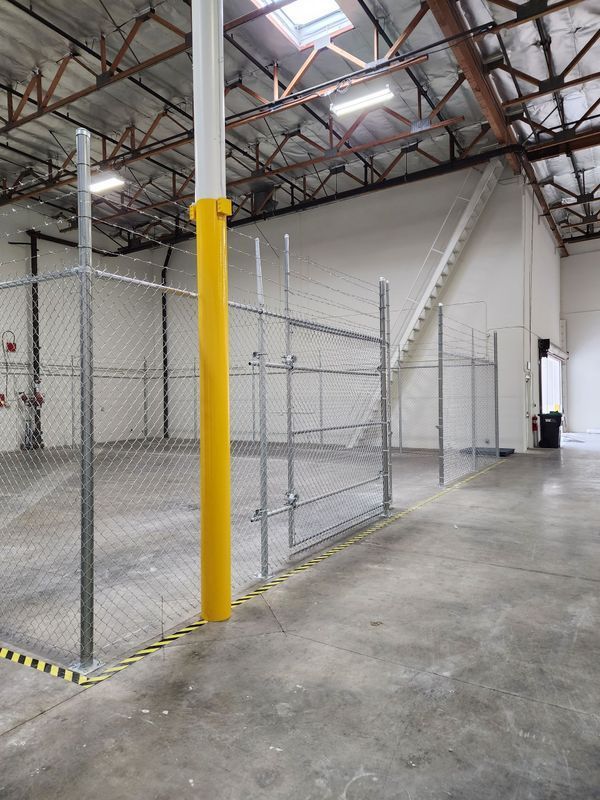 A chain-link fence inside an industrial warehouse, yellow pole protector, stairway in background.