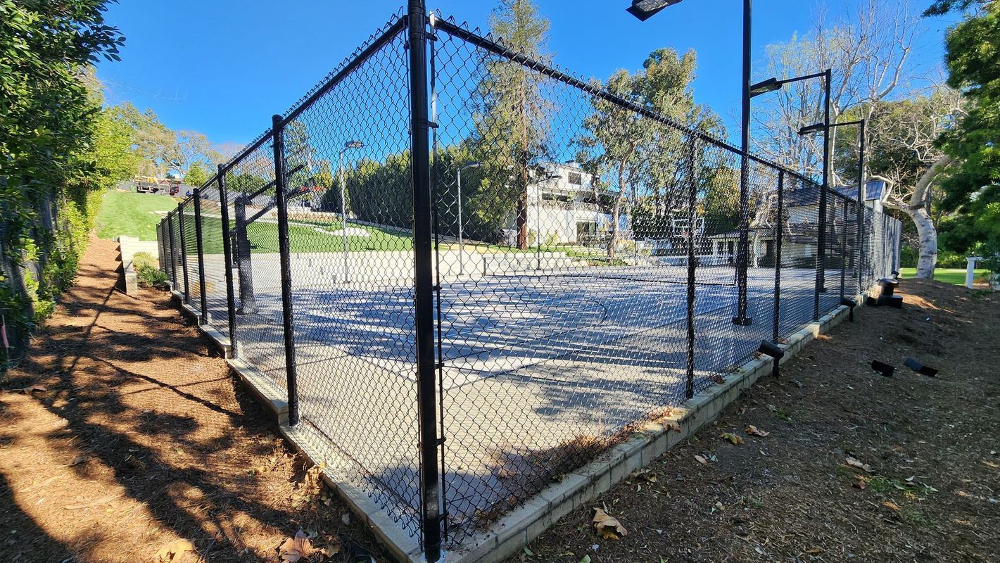 Tennis court enclosed by black fence, trees in background. Blue sky.